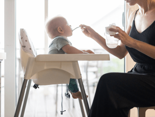 mãe dando comida a um bebê que está sentado em uma cadeira de alimentação,seguindo os 5 Princípios Básicos da Introdução Alimentar