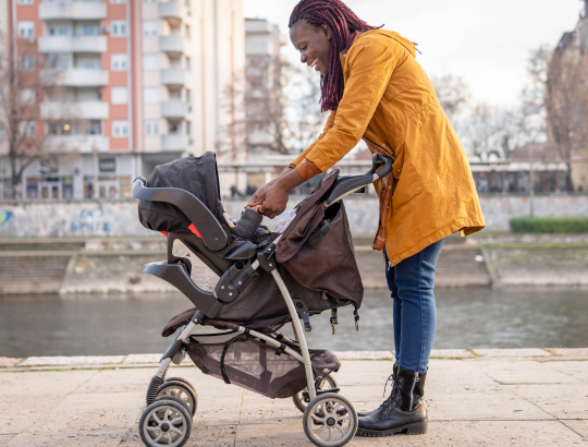 Mulher preta vestindo um casaco amarelo segurando as mãos de um bebê que está dentro de um carrinho travel system reverse da Cosco é bom