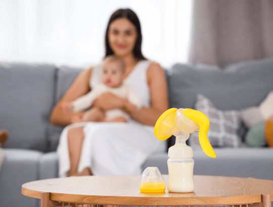 um dos melhores coletores de leite materno sobre uma mesa e ao fundo, uma mãe com um bebê no colo
